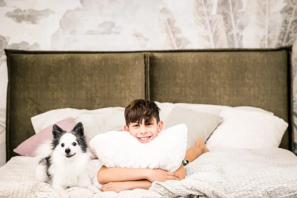 Junge liegt in einem modernen Boxspringbett und stützt seinen Kopf auf ein Kissen, neben ihm liegt ein Hund